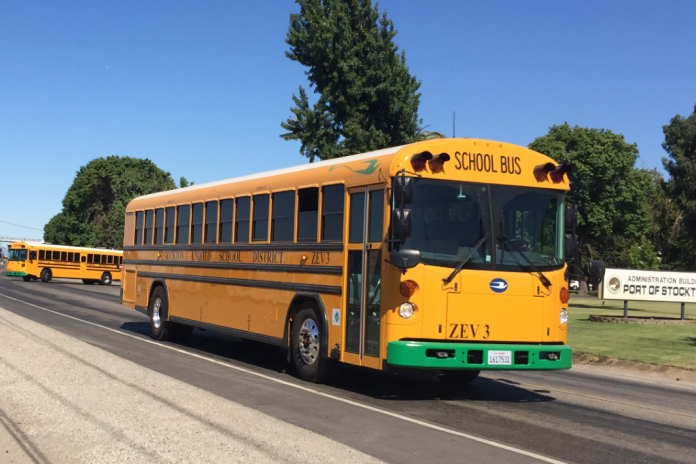 A yellow electric school bus drives down a road, as another pulls out onto the same road behind it. A sign beside the road reads: "Administration Building: Port of Stockton".