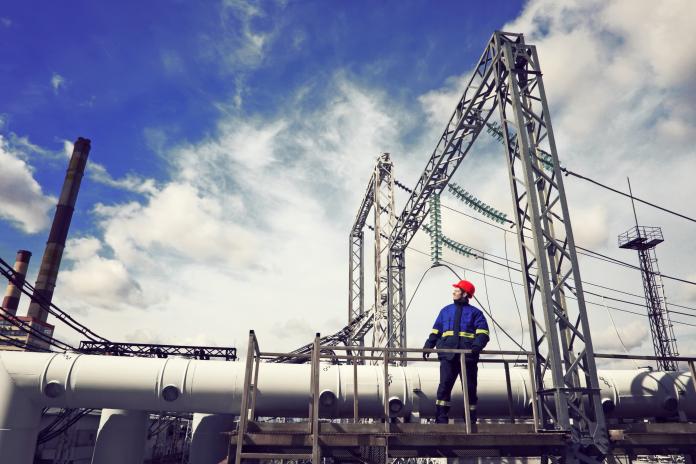 A worker stands atop an electrical power plant. There are blue skies and clouds in the background.