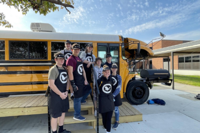 A group of students wearing hats and aprons stand outside an old school bus, which has been repurposed into a coffee shop.