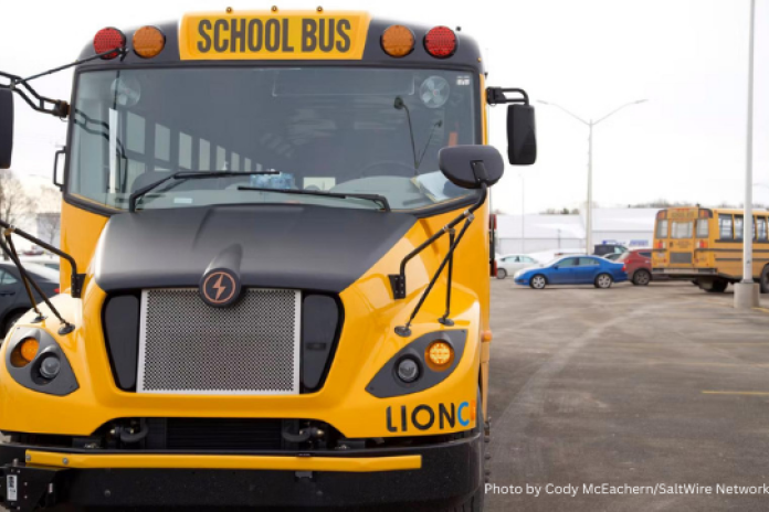 A picture of a yellow electric school bus, taken from the front. In the background is another school bus and some cars.