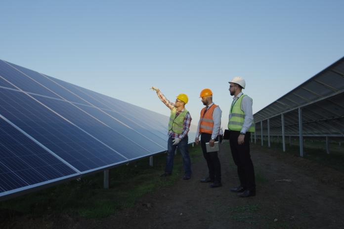Three workers in hard hats stand near a solar panel and point toward the sky.