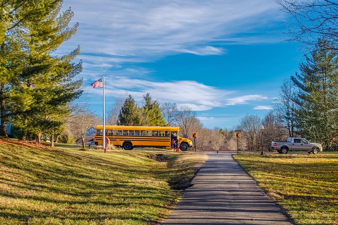 A yellow school bus at a bus stop with blue skies, trees and American flag on a pole nearby.