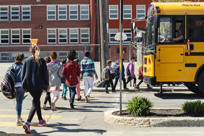 Students are lined up, walked away from the camera to board a yellow school bus, in front of a school.