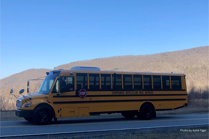 Yellow electric school bus with the words "Cherokee Boys Club Bus Service" written on the side drives on a two-lane road under a blue sky in front of brown mountains.