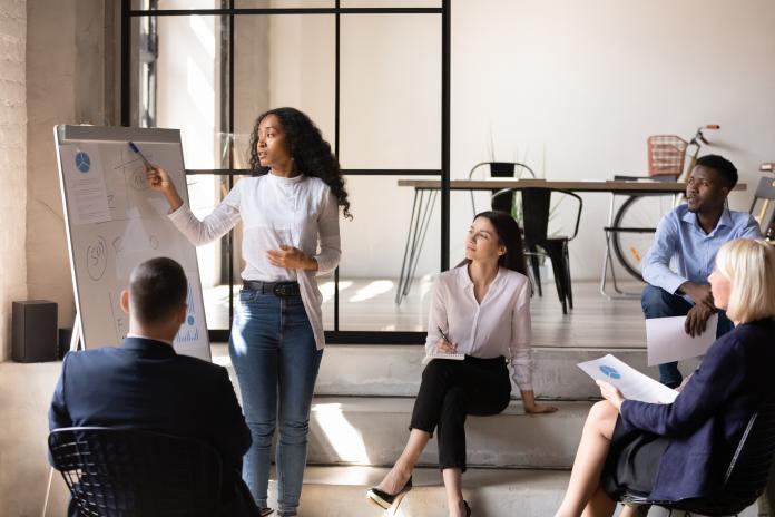 A woman stands in a meeting room at a white board and points while a group of people in business casual attire look on.