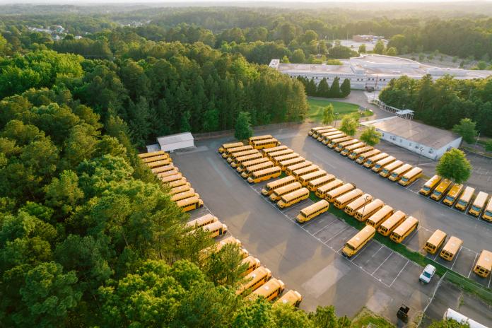 Several dozen yellow school buses are parked in a parking lot, surrounded by trees and seen from above.