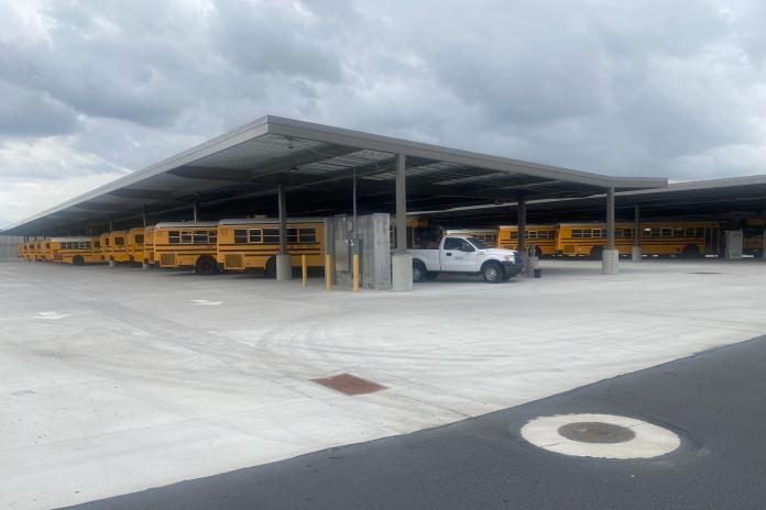 Electric school buses charge in a parking lot, under a canopy.