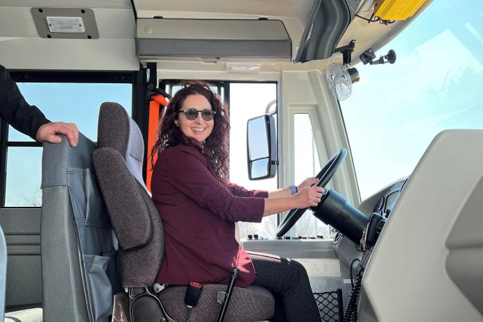 A person wearing sunglasses and a seat belt sits at the wheel of an electric school bus.