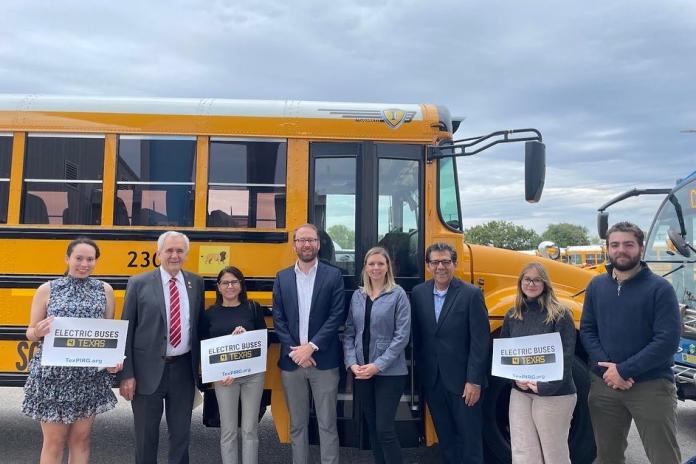 Electric school bus advocates and lawmakers stand in front of a school bus, holding signs that read "Electric Buses 4 Texas"