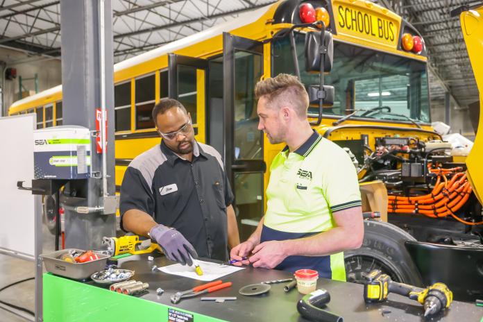 Electric school bus technicians work at a table near a school bus.