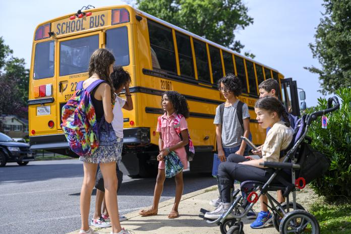 A group of students, including a student in a wheelchair, talk to one another in front of a parked yellow electric school bus.