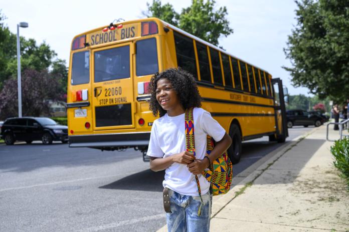 A student holds a backpack near with an electric school bus on the background.