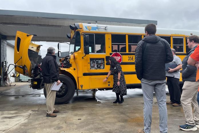 A group of people stand in front of an electric school bus with its hood open