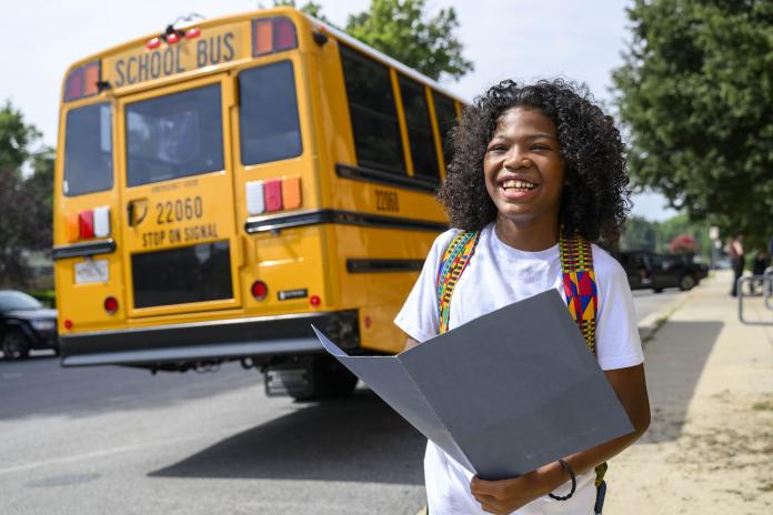 A student holds a folder near an electric school bus and smiles.
