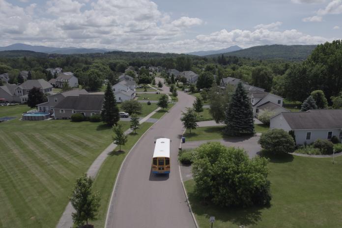 An electric school bus driving through a tree-lined residential neighborhood with mountain views in the background, photographed from an aerial perspective.