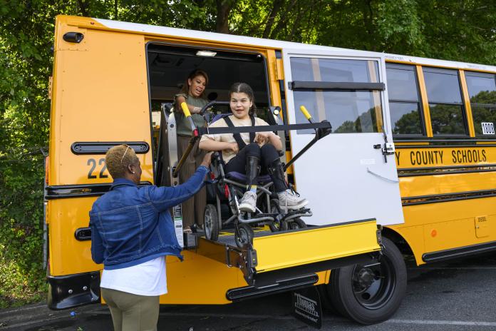 A student uses a wheelchair lift to deboard a school bus with the help of two adults.