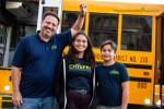 Three people stand in front of an electric school bus smiling as one raises his fist.