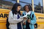 Students look at a textbook while standing near a school bus.