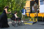 A student using a wheelchair boards a school bus wheelchair lift, with multiple aides and adults assisting.