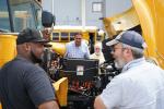 Two people stand and speak near an open electric school bus hood.