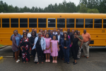 A group of about 25 people of various ages stand and smile in front of an electric school bus.