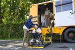 A student in a wheelchair deboards an electric school bus using a wheelchair lift, with help from the driver
