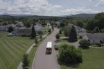 An electric school bus driving through a tree-lined residential neighborhood with mountain views in the background, photographed from an aerial perspective.