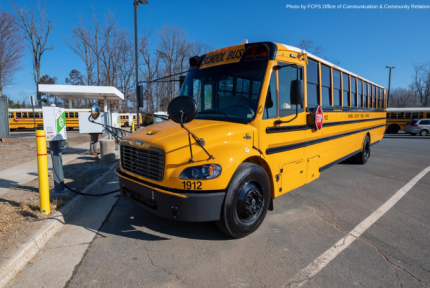 A yellow electric school bus is parked in a bus lot and is plugged in, charging.