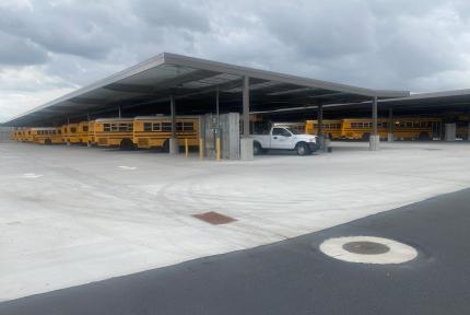 Electric school buses charge in a parking lot, under a canopy.