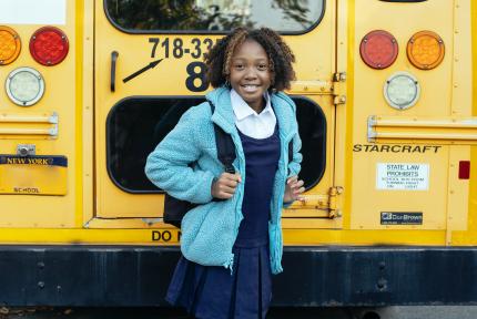 A child in a backpack stands smiling behind a school bus.