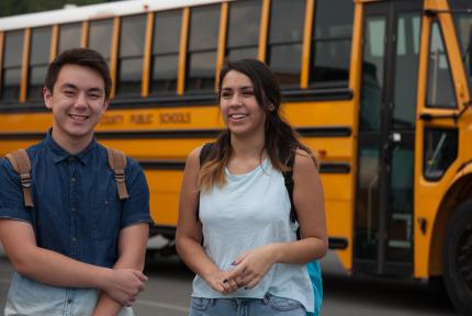 Students smile while wearing backpacks near a school bus.