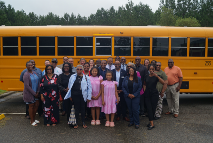 A group of about 25 people of various ages stand and smile in front of an electric school bus.