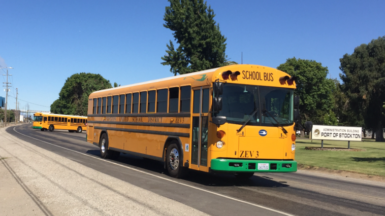 A yellow electric school bus drives down a road, as another pulls out onto the same road behind it. A sign beside the road reads: "Administration Building: Port of Stockton".