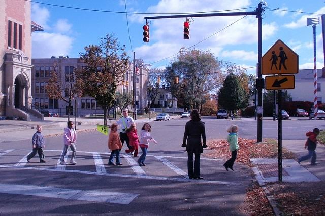 Young students walk across a crosswalk at a red light, accompanied by a crossing guard and another adult.