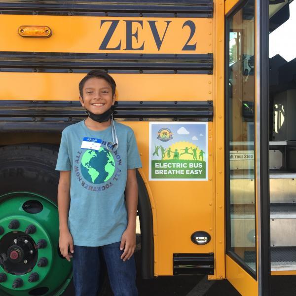 A young, smiling boy stands outside of the entrance to a yellow electric school bus. Near the door, a flyer is posted that reads: "Electric Bus Breathe Easy".