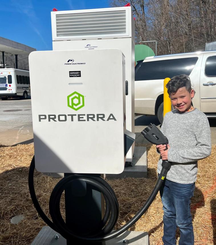 Young boy stands holding an electric bus charger plug. The charger itself reads "Proterra".