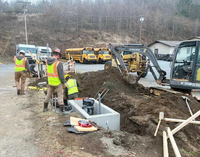Men in construction outfits dig a large hole to install electric bus chargers. They are surrounded by heavy machinery. School buses seen in the background.