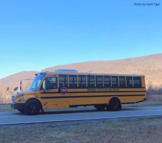 A photo of the side of a yellow electric school bus, driving on the road. Text on bus reads "Cherokee Boys Club Bus Service". Mountains and blue sky in the background.