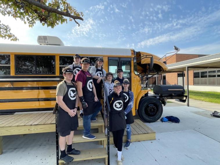 Student wearing aprons and hats pose outside of an old school bus, which has been transformed into a coffee shop. The bus is located in front of a school.