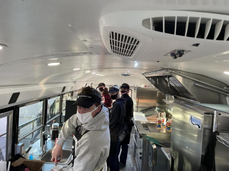 Photo inside of an old school bus, which has been transformed into a coffee shop. 4 students in aprons and hats and masks look at the camera.