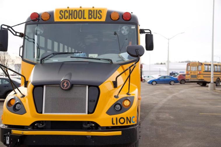 A picture of a yellow electric school bus taken from the front. On the right side of the photo in the background is another school bus and some more cars.