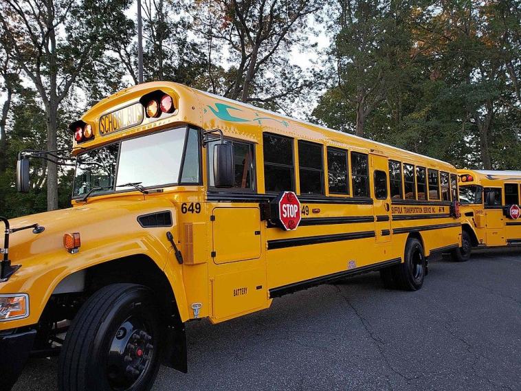 Photo of an electric school bus from the front/side. Trees are behind the bus, and another bus is parked behind.