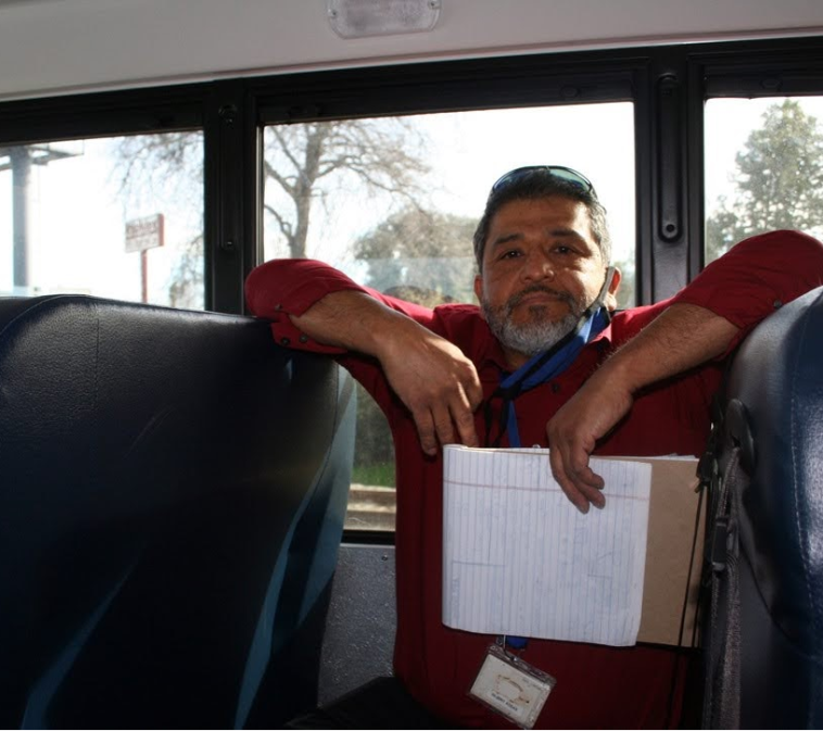 A man, Gilbert Rosas, sits in one of the seats of an electric school bus with arms resting on the tops of the seats. He holds a notepad.