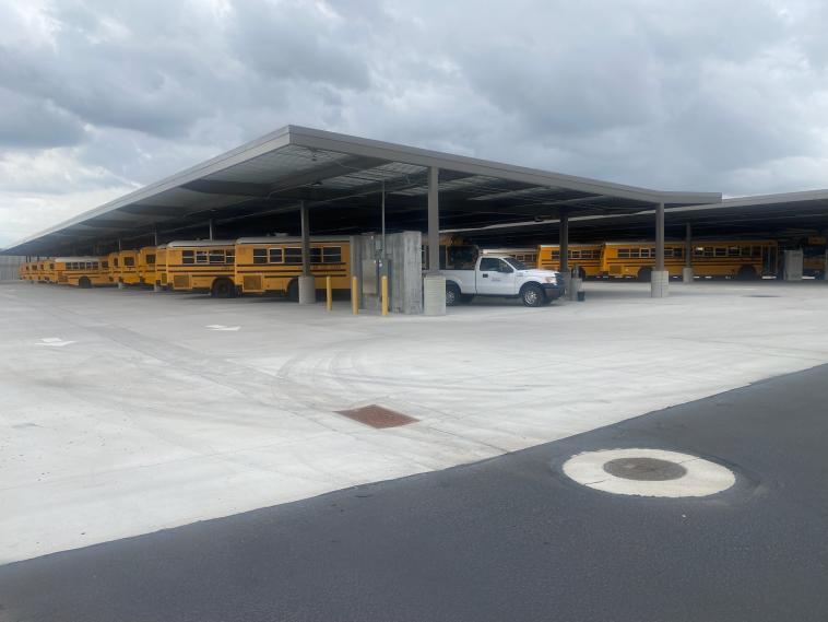 Electric school buses charge in a parking lot, under a canopy.