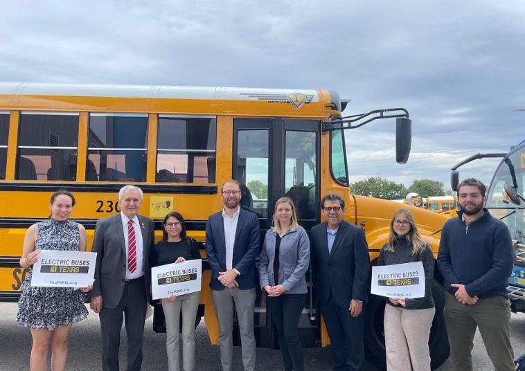 Electric school bus advocates and lawmakers stand in front of a school bus, holding signs that read "Electric Buses 4 Texas"