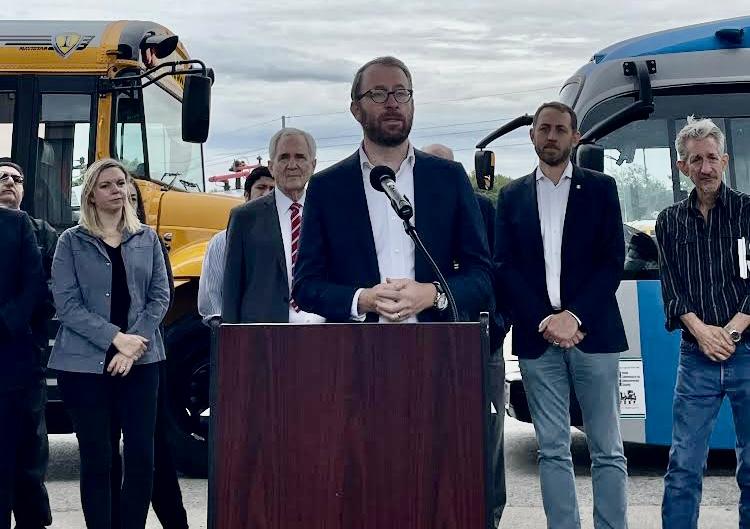 A man stands at a lectern speaking, with a group of people and two buses behind him.