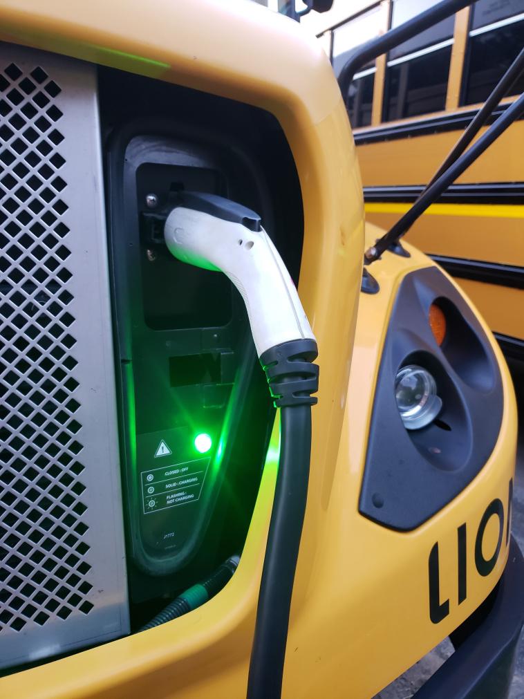 A close-up shot of a white charger plugged into a yellow school bus. A green light is on.