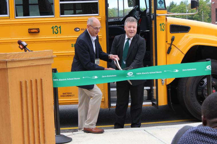 Two people stand in front of an electric school bus, cutting a green ribbon with large scissors.