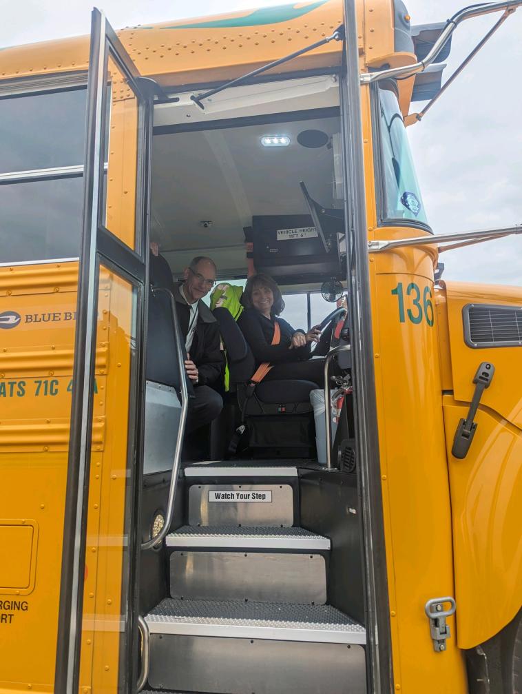 Two people smile near the driver’s seat of an electric school bus.