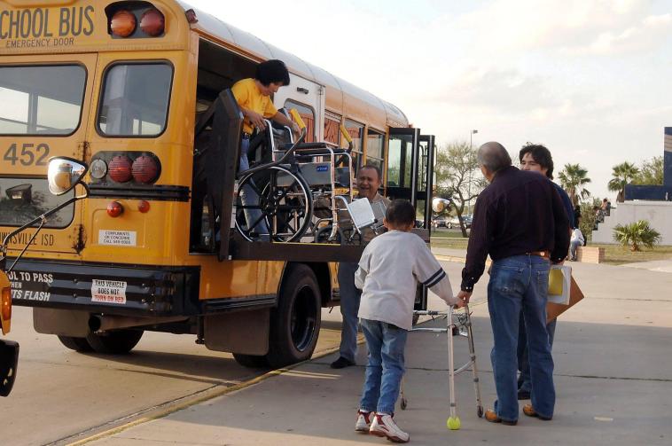 A student using a wheelchair boards a school bus using a wheelchair lift, while a student using a walker waits. Adults and aides assist.